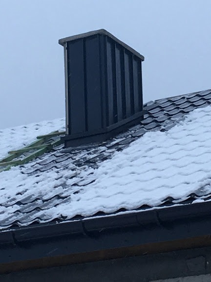 Metalowy komin na dachu pokrytym śniegiem i dachówką ceramiczną, widoczne elementy konstrukcji drewnianej, pochmurne niebo w tle.