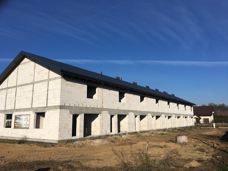 Budowa szeregowca z białych bloczków w Głogowie koło Torunia, z zamontowanym ciemnym dachem, widoczne niezabudowane otwory okienne i drzwiowe, oraz tablica reklamowa osiedla.