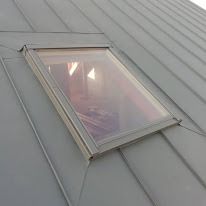 Szary dach z blachy na rąbek stojący z oknem dachowym; widok z dołu pod kątem.