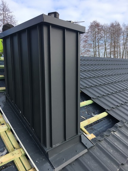 Nowy, czarny komin z blachy na dachu krytym dachówką bitumiczną, widoczne deski łat dachowych i fragment roboczej odzieży dekarza.