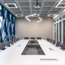 Nowoczesna sala konferencyjna z dużym stołem w kształcie litery U, szarymi krzesłami i geometrycznym oknem z widokiem na miasto, oświetlona kwadratowymi lampami LED i podłużną lampą w kształcie...