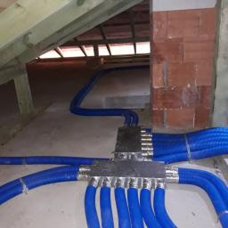 Ekopompaciepła.pl Sp. z o.o. - Rozdzielacz z podłączonymi niebieskimi rurami wentylacyjnymi na poddaszu budynku w Skrzeszewie, widoczne elementy konstrukcji dachu i komin z cegły.