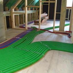 Ekopompaciepła.pl Sp. z o.o. - Wnętrze w trakcie budowy z widocznym fragmentem instalacji ogrzewania podłogowego: rury w kolorze zielonym i czerwono-niebieskim ułożone na surowej drewnianej podłodze, przymocowane metalowymi...