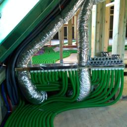 Ekopompaciepła.pl Sp. z o.o. - System wentylacyjny z zielonymi rurami i srebrnymi kanałami w surowym wnętrzu poddasza, widoczne drewniane elementy konstrukcyjne i okno dachowe.