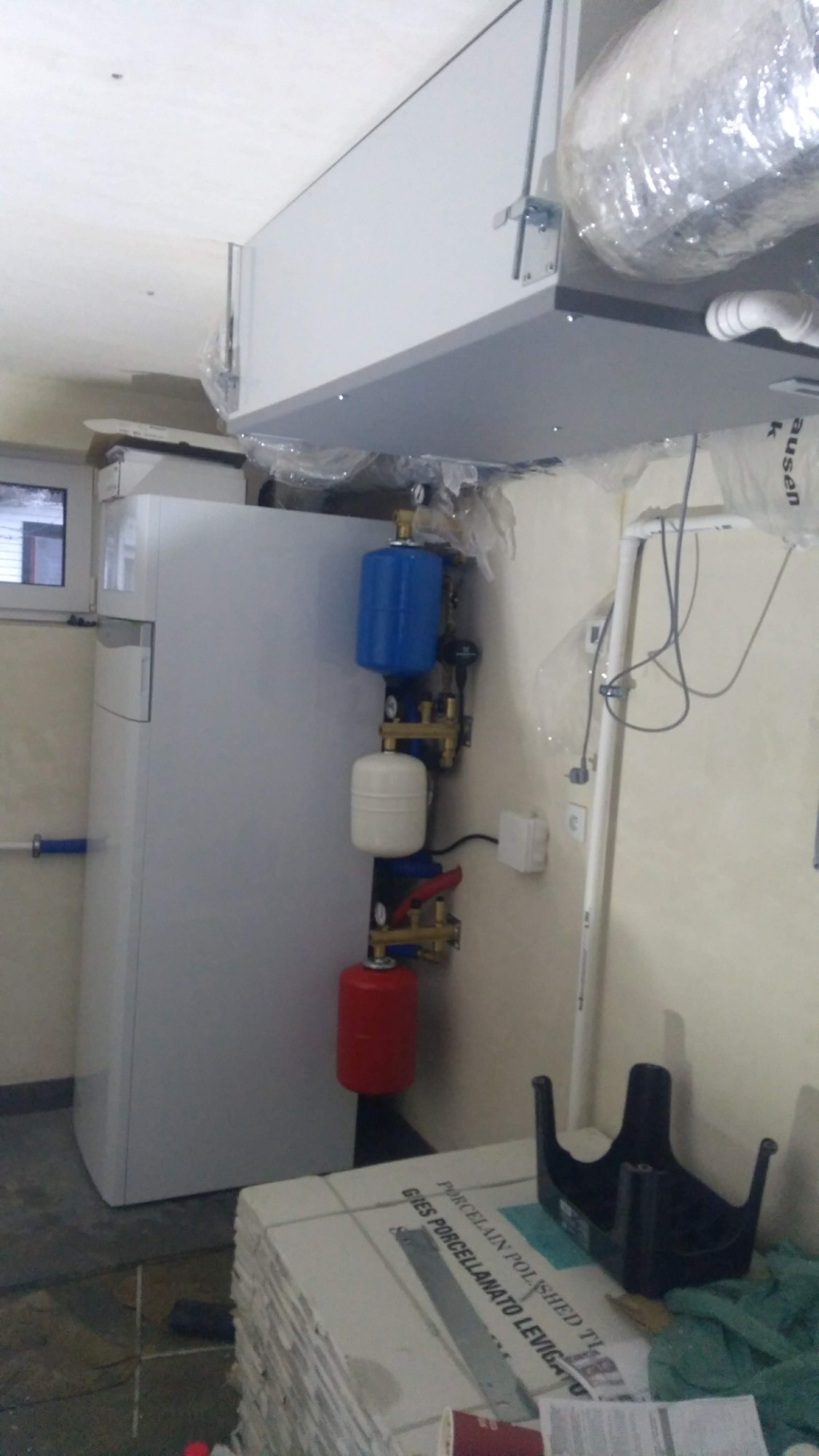 Wnętrze kotłowni z zamontowaną jednostką centralną, prawdopodobnie pompą ciepła, widoczne zbiorniki wyrównawcze w kolorach niebieskim, białym i czerwonym, instalacja pod sufitem oraz sterta płytek...