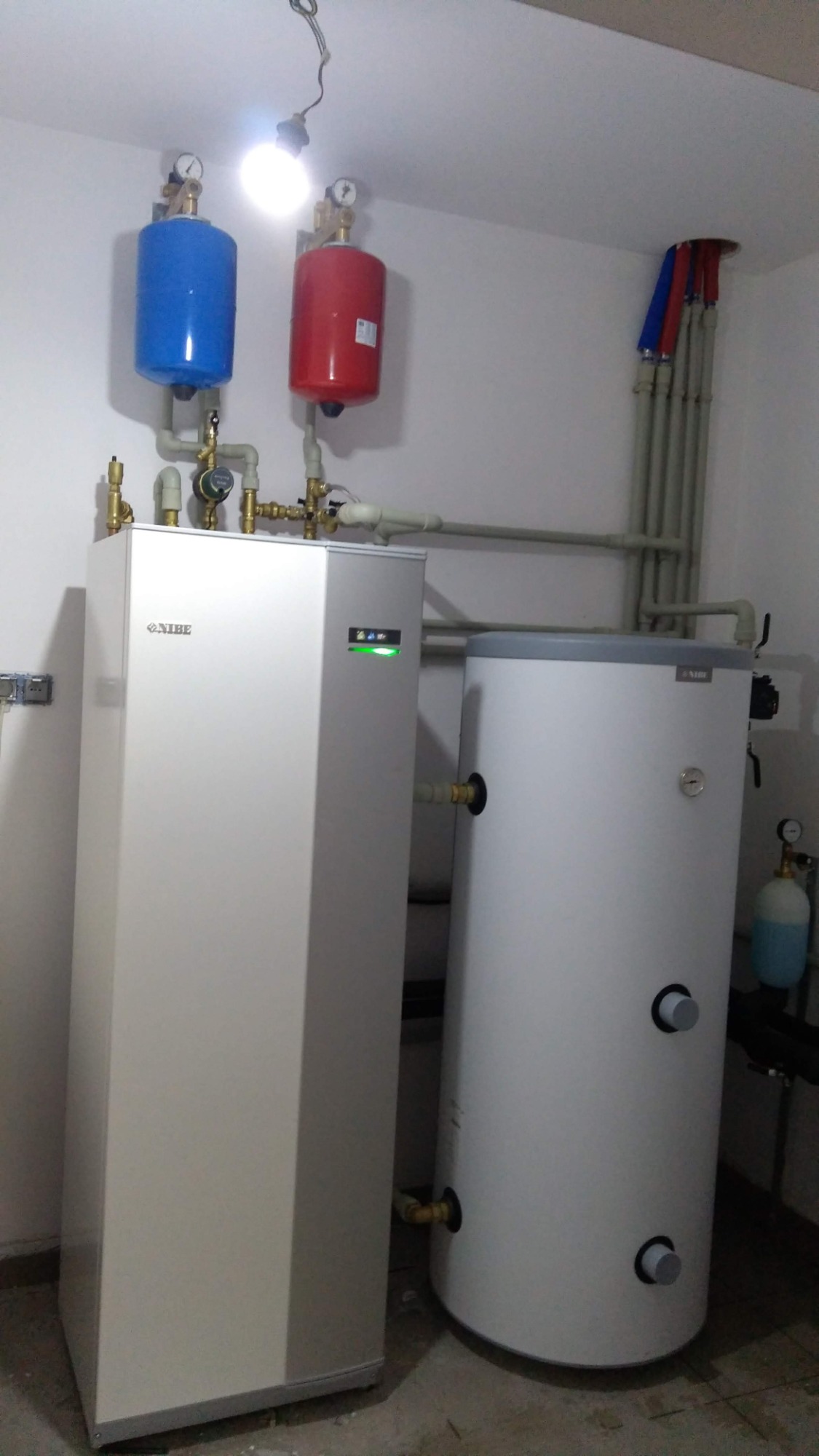 Biała jednostka wewnętrzna z logo Nibe, podłączona do instalacji z czerwonym i niebieskim zbiornikiem wyrównawczym, oraz biały zasobnik ciepłej wody w kotłowni z widocznymi rurami.