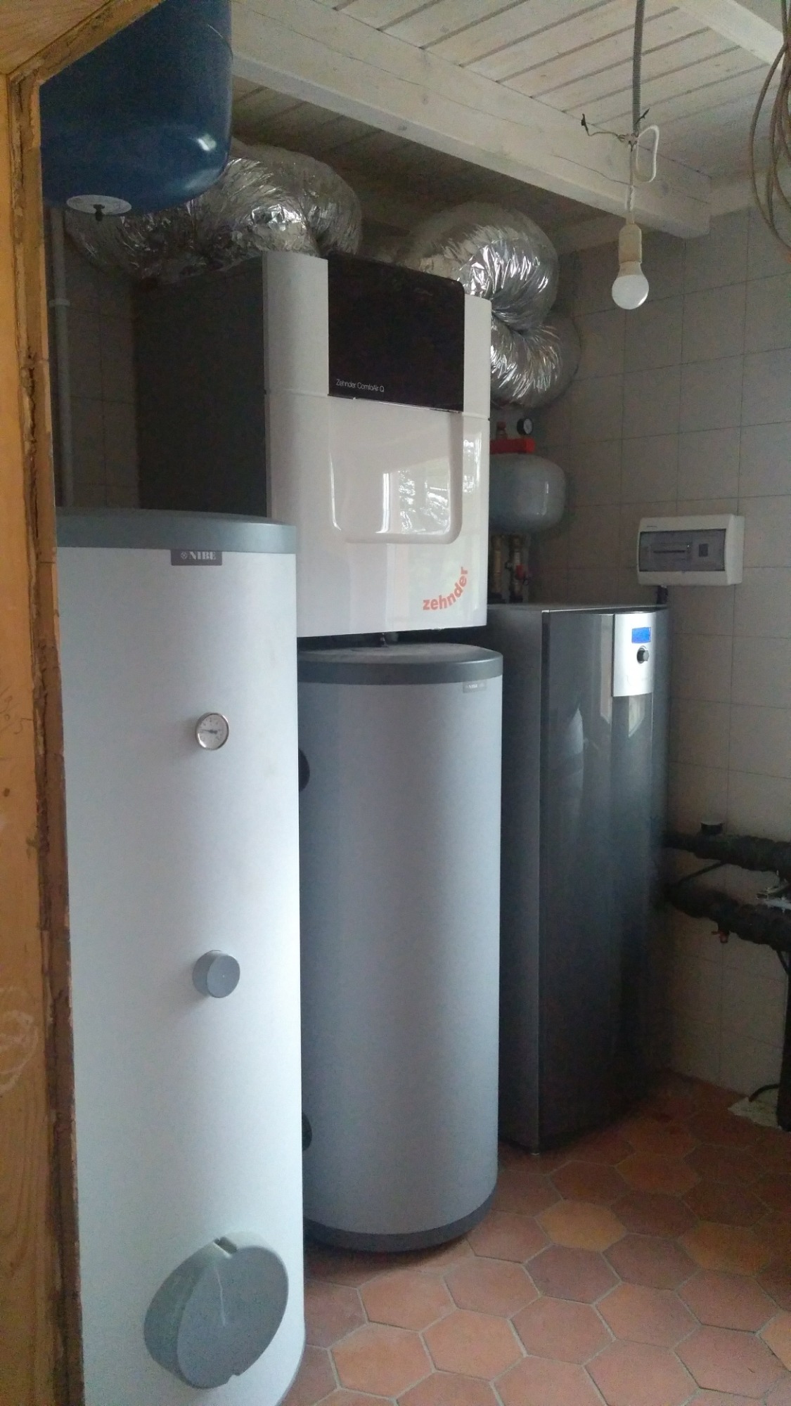 Zestaw urządzeń grzewczych w kotłowni: dwa białe zasobniki Nibe z szarymi akcentami, centrala wentylacyjna Zehnder Comfo Q z czarnym panelem, oraz srebrne rury wentylacyjne pod sufitem, na tle...