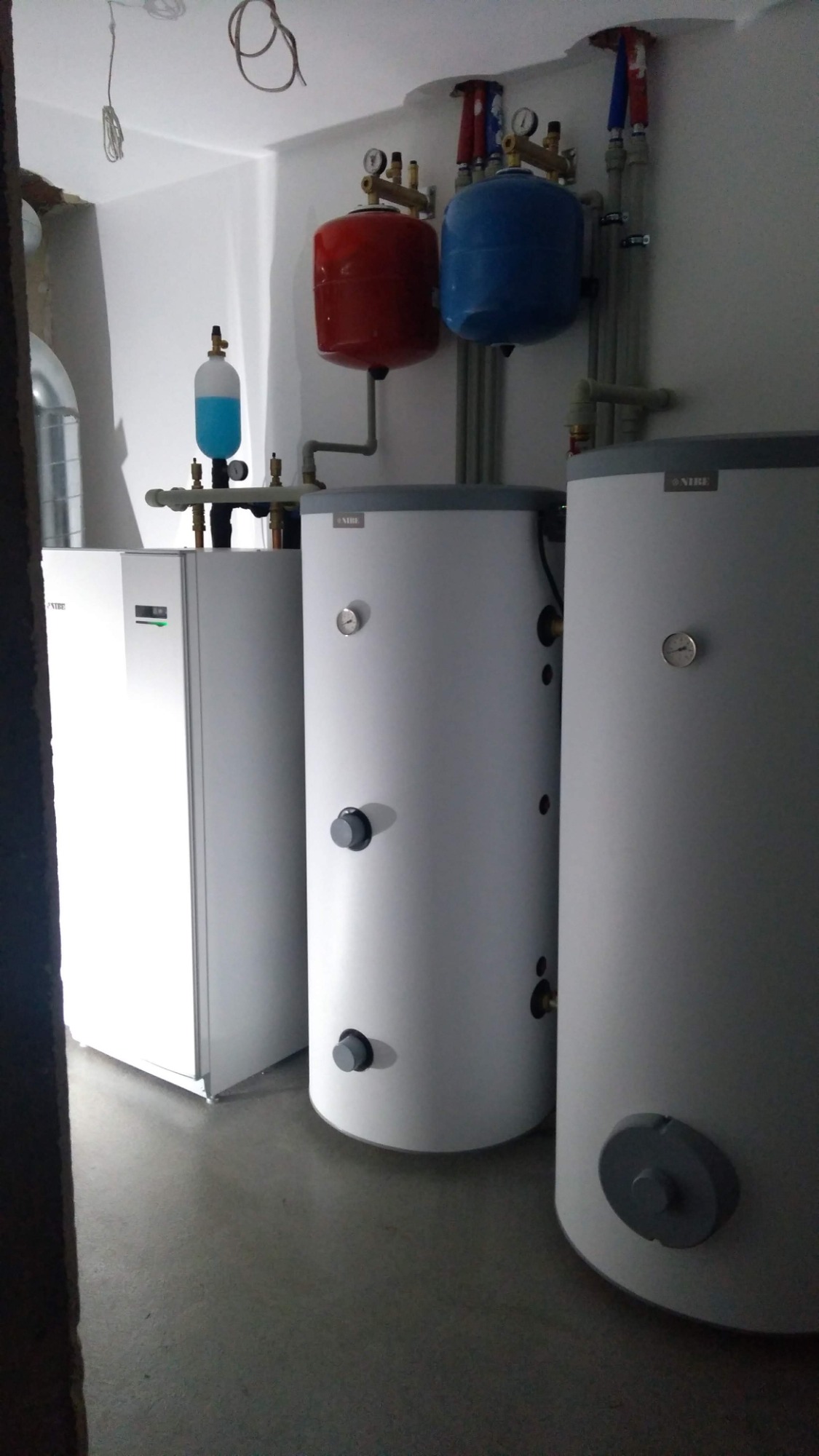 Instalacja grzewcza w pomieszczeniu: biała jednostka, dwa białe zbiorniki z szarymi elementami, czerwony i niebieski zbiornik wyrównawczy, rury miedziane i plastikowe, manometry, zawory, instalacja...