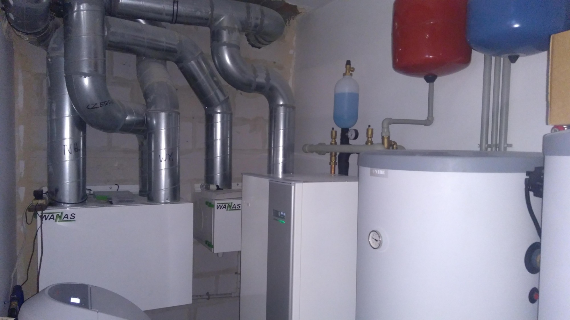 Instalacja pompy ciepła WANAS z widocznymi rurami wentylacyjnymi, zbiornikiem wyrównawczym i zasobnikiem ciepłej wody w surowym pomieszczeniu z nieotynkowanymi ścianami.