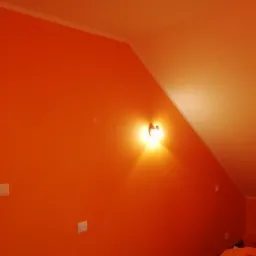 Pomarańczowa ściana w pokoju na poddaszu, oświetlona kinkietem. Widoczne białe włączniki światła i fragment brązowej futryny drzwi.