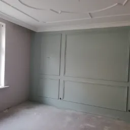 Pokój po remoncie z pastelowymi ścianami, sztukaterią i listwami przypodłogowymi, widok z okna na zewnątrz, podłoga zabezpieczona folią.
