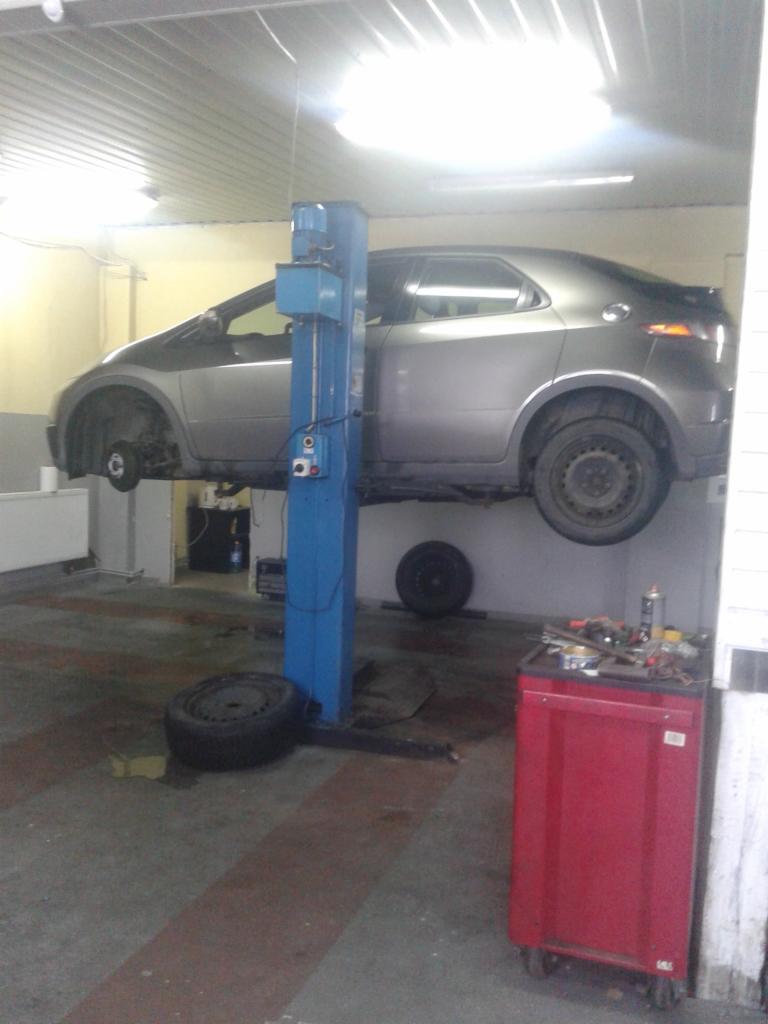 Szary samochód na podnośniku w warsztacie, brakujące koła, widoczny wózek narzędziowy i opona na podłodze.