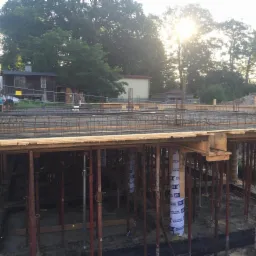 Konstrukcja zbrojeniowa płyty fundamentowej wsparta na metalowych stemplach, widoczne deskowanie i przygotowanie do betonowania, w tle budynki i drzewa w letnim słońcu.