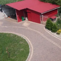 Szeroki podjazd z kostki brukowej w odcieniach brązu, z dekoracyjnym motywem liścia z jaśniejszej kostki, prowadzący do czerwonego budynku gospodarczego z otwartą wiatą garażową, otoczonego zielenią.