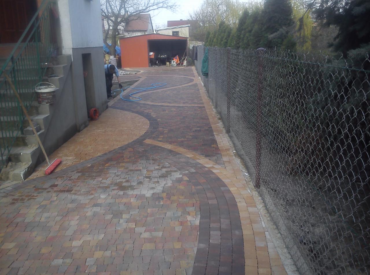 Umyta wodą, kolorowa kostka brukowa ułożona w dekoracyjny wzór na podwórku, widoczny pracownik z wężem, sąsiadująca siatka ogrodzeniowa i budynek gospodarczy w tle.