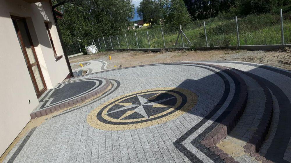 Ułożona nawierzchnia z kostki brukowej z dekoracyjnym motywem rozety w kolorach szarości, czerni i żółci, obrzeża z kostki w kolorze ceglastym, widok na dom i ogrodzenie.