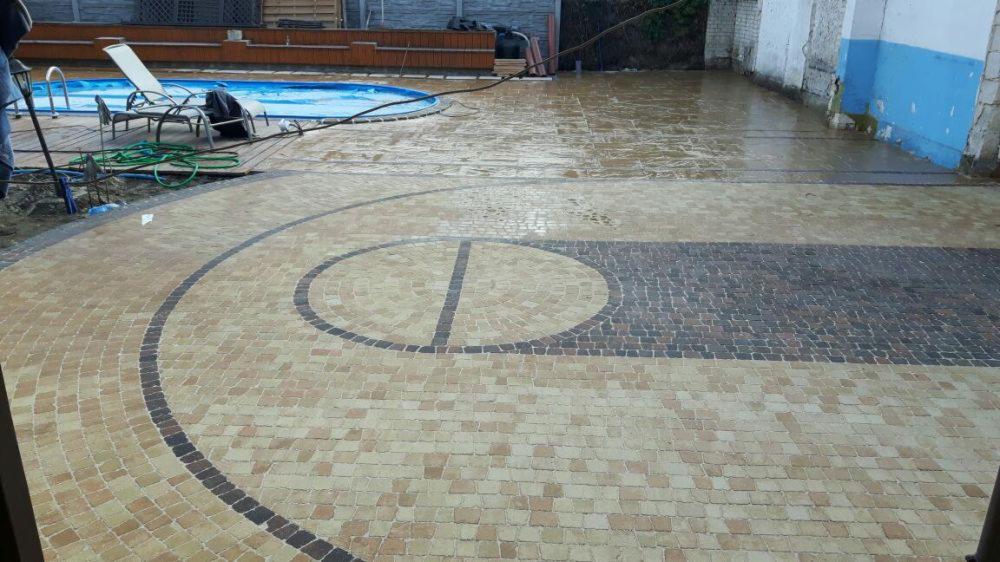 Ułożona nawierzchnia z kostki brukowej w odcieniach beżu i szarości, z dekoracyjnym wzorem koła i linii, otoczona basenem i elementami drewnianej architektury ogrodowej.