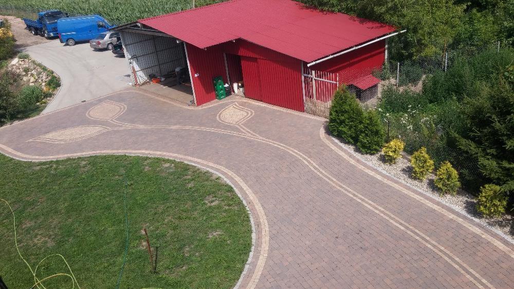 Szeroki podjazd z kostki brukowej w odcieniach brązu, z dekoracyjnym motywem liścia z jaśniejszej kostki, prowadzący do czerwonego budynku gospodarczego z otwartą wiatą garażową, otoczonego zielenią.