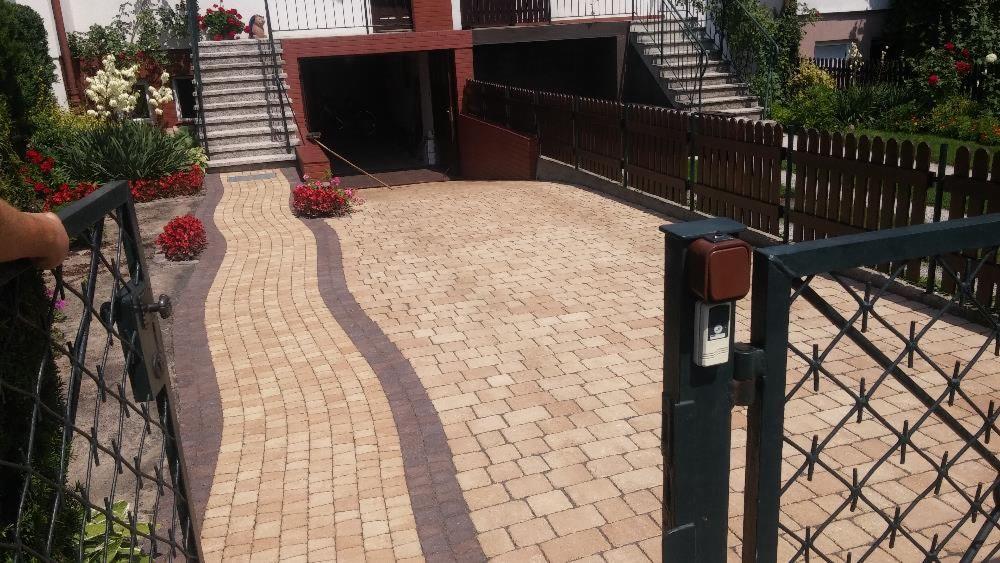 Ułożona beżowa kostka brukowa z ciemniejszymi pasami tworzącymi falisty wzór na podjeździe, widok zza otwartej metalowej furtki, w tle budynek z garażem i schodami, zadbana roślinność.