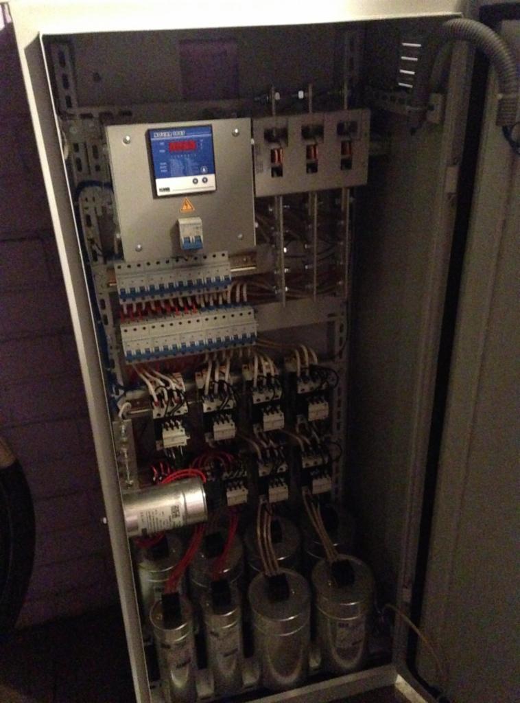 Wnętrze szafy sterowniczej z widocznymi stycznikami, bezpiecznikami, kondensatorami i przewodami, zamontowanej prawdopodobnie przy bramie.