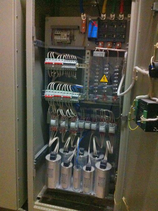 Otwarta szafa sterownicza z kondensatorami, bezpiecznikami i okablowaniem, widoczny znak ostrzegawczy wysokiego napięcia.