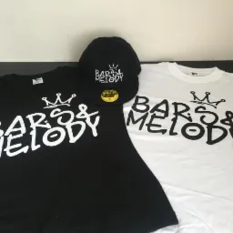 Dwie koszulki (czarna i biała) oraz czapka z daszkiem, wszystkie z białym nadrukiem 'Bars & Melody' z motywem korony, ułożone obok siebie na ciemnym blacie.