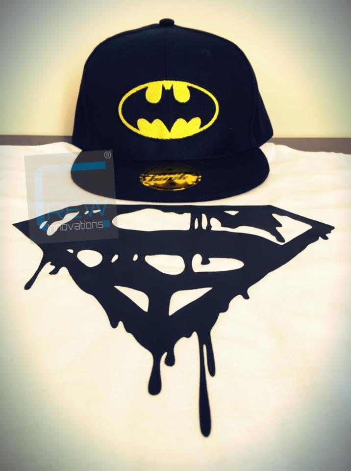 Czapka z daszkiem w kolorze granatowym z żółtym logo Batmana, leżąca obok czarnego szablonu z motywem Supermana, imitującego efekt topienia się.