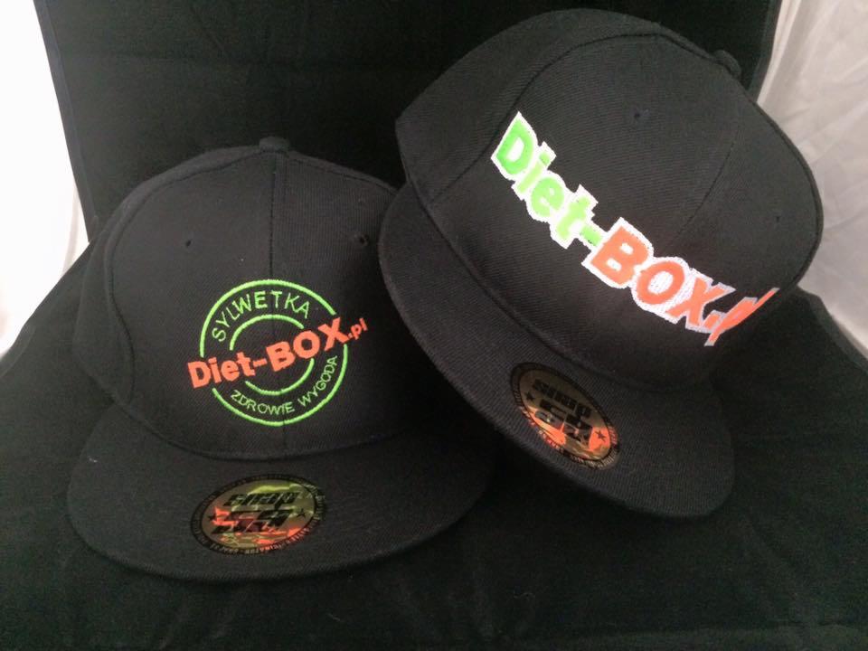 Dwie czarne czapki typu snapback z kolorowymi logotypami 'Diet-BOX' i 'SYLWETKA Diet-BOX.pl', leżące na ciemnym, miękkim tle.