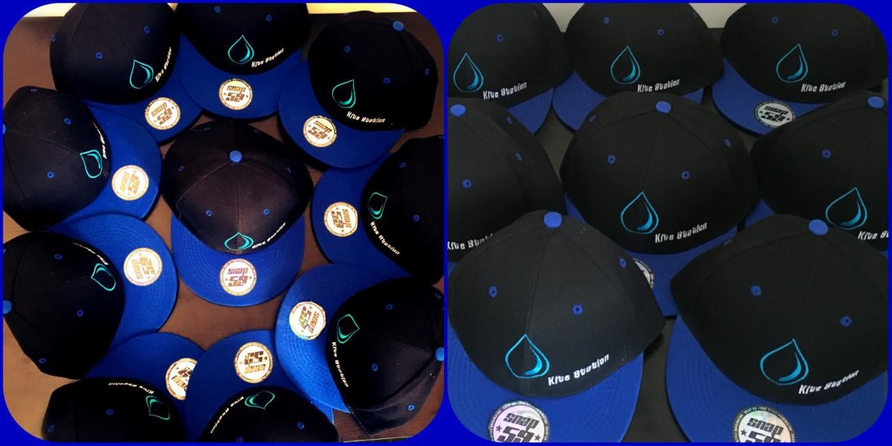 Czapki z daszkiem w kolorze czarnym i niebieskim z wyhaftowanym logo firmy z Kołobrzegu, ułożone w stos. Widoczne detale haftu i naszywki.