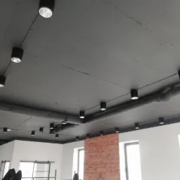 Równomiernie rozmieszczone, czarne reflektory punktowe na szarym suficie w nowoczesnym wnętrzu z fragmentem ściany z czerwonej cegły i instalacją wentylacyjną.