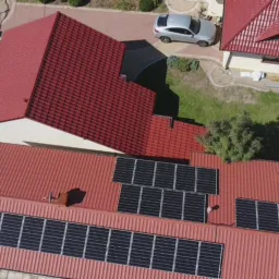 Widok z góry na dom z czerwonym dachem i zainstalowanymi panelami słonecznymi; obok zaparkowany srebrny samochód.