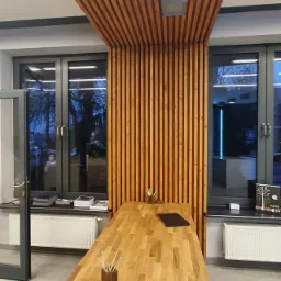 Nowoczesne wnętrze z drewnianą zabudową sufitu i ściany, drewniany stół w centrum, widok na okna i grzejniki.