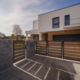 Nowoczesny dom z drewnianą elewacją i przesuwną bramą z kompozytowych desek, numer domu '53' wyryty w betonowym murku, jasna kostka brukowa na podjeździe.