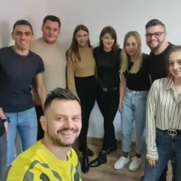 Grupa uśmiechniętych osób, w tym mężczyzna w żółtej koszulce z przodu robiący selfie, prawdopodobnie po szkoleniu firmowym.