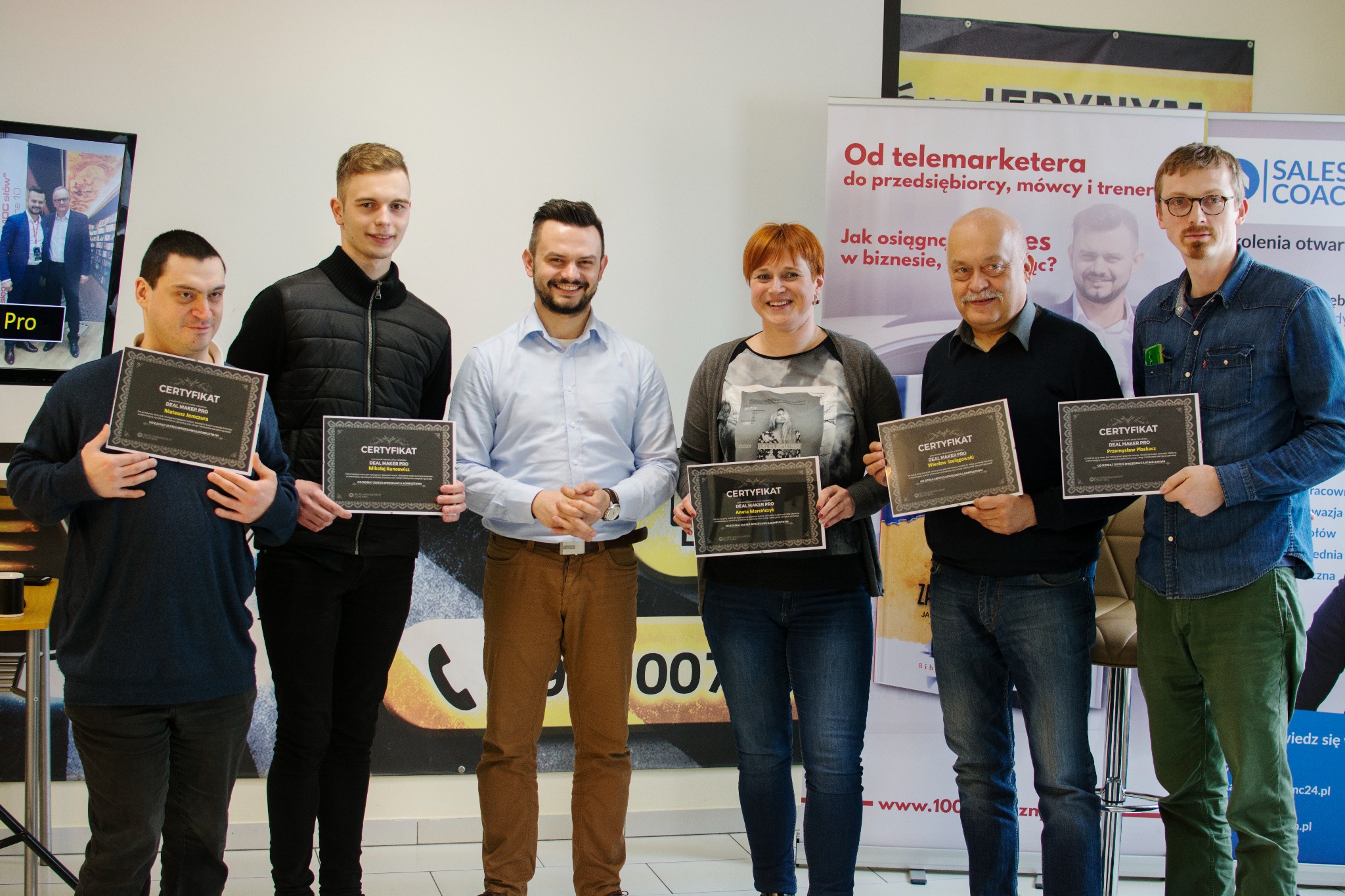 Grupa sześciu osób, w tym jedna kobieta, prezentuje certyfikaty ukończenia szkolenia DEAL MAKER PRO, stojąc na tle grafiki reklamowej z hasłem 'Od telemarketera do przedsiębiorcy, mówcy i trenera'.