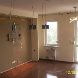 Wnętrze pokoju po remoncie, z zamontowanym uchwytem na telewizor, wiszącymi lampami i zabezpieczonym grzejnikiem.