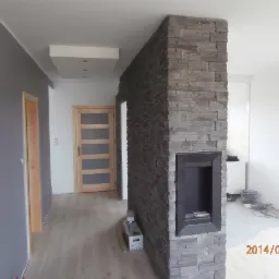 Nowoczesny kominek z obudową z szarego kamienia dekoracyjnego, wbudowany w ścianę działową między pokojami, widok na salon i korytarz z drewnianymi drzwiami.