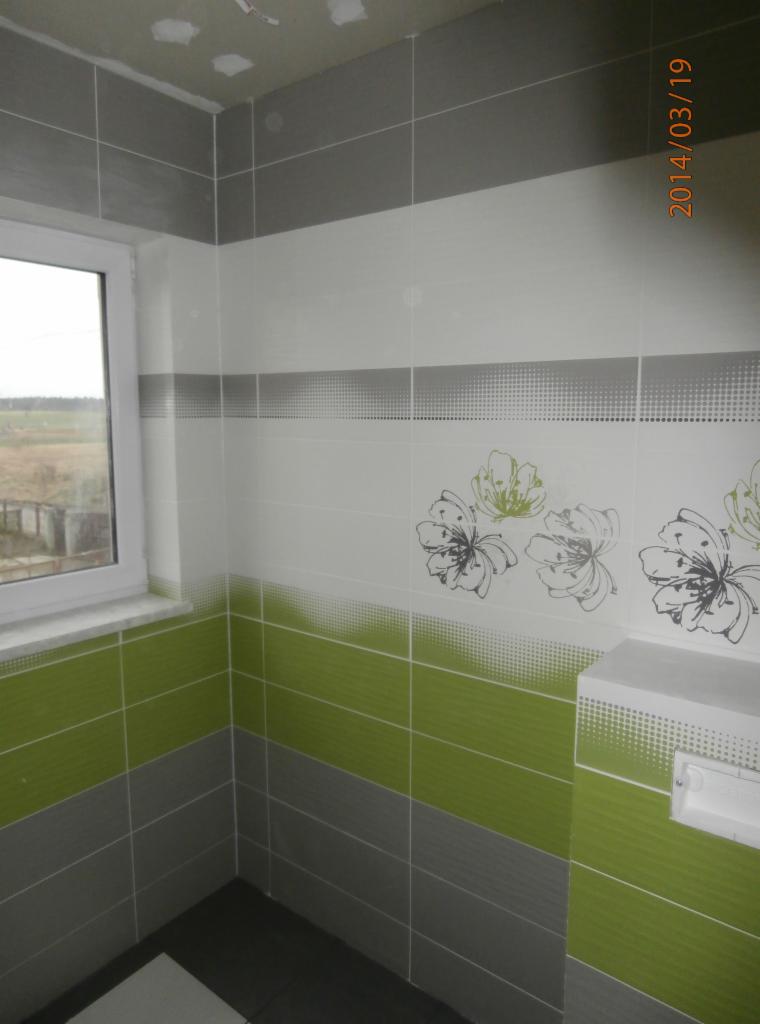 Świeżo wykafelkowana ściana łazienki z motywem kwiatowym i paskami w odcieniach szarości i zieleni, widoczne okno z widokiem na krajobraz.