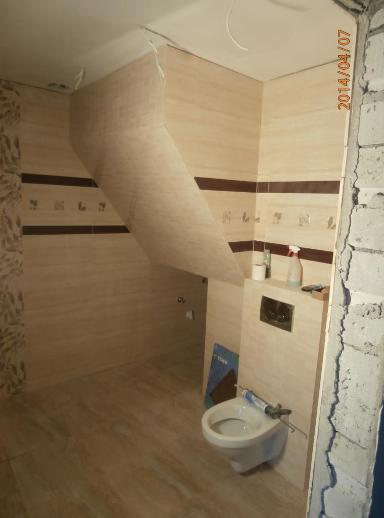 Niedokończona łazienka z beżowymi płytkami, widoczne narzędzia i nieotynkowana ściana, umywalka podwieszana i częściowo zabudowana instalacja sanitarna.