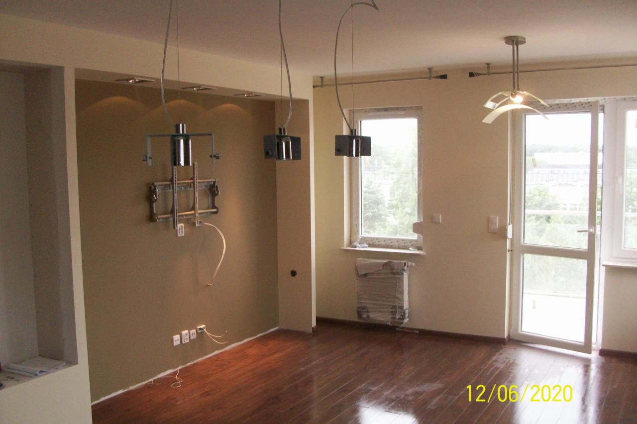 Wnętrze pokoju po remoncie, z zamontowanym uchwytem na telewizor, wiszącymi lampami i zabezpieczonym grzejnikiem.