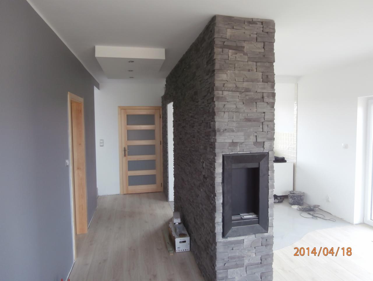 Nowoczesny kominek z obudową z szarego kamienia dekoracyjnego, wbudowany w ścianę działową między pokojami, widok na salon i korytarz z drewnianymi drzwiami.
