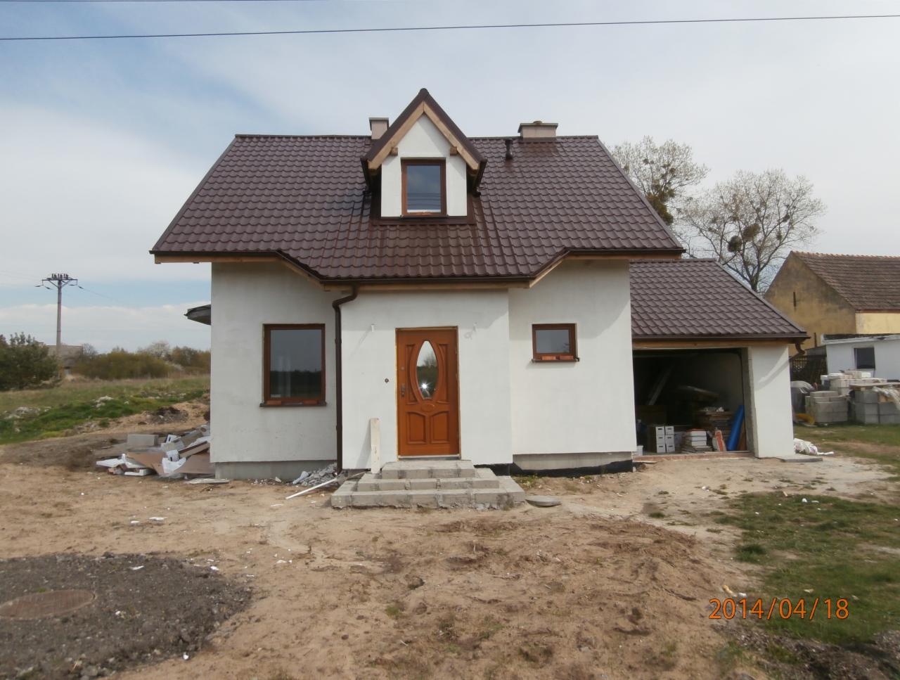 Biały dom jednorodzinny z brązowym dachem z dachówki, drewnianymi oknami i drzwiami wejściowymi, widok z przodu na plac budowy z widocznymi materiałami budowlanymi.