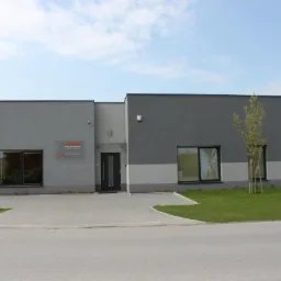 Nowoczesny budynek biurowy w odcieniach szarości z logo firmy na elewacji, otoczony trawnikiem i młodym drzewkiem.