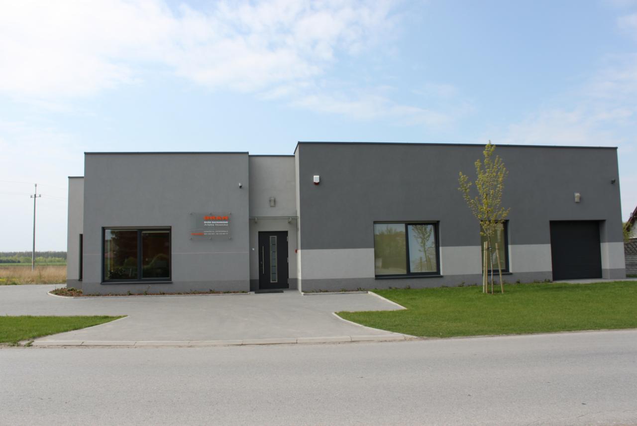 Nowoczesny budynek biurowy w odcieniach szarości z logo firmy na elewacji, otoczony trawnikiem i młodym drzewkiem.