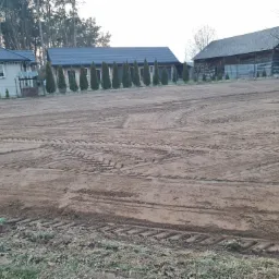 Przygotowany teren pod budowę z widocznymi śladami opon maszyn budowlanych, w tle budynki mieszkalne i gospodarcze otoczone drzewami.