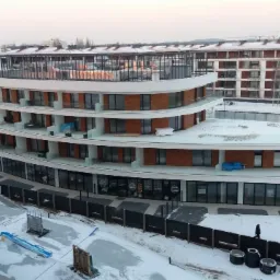 Wielopiętrowy budynek mieszkalny z balkonami i szklaną nadbudówką w zimowej scenerii, widok z góry z fragmentem żurawia budowlanego w kadrze.