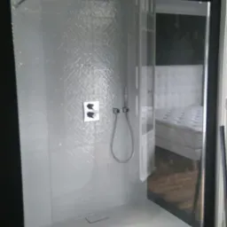 Nowoczesna kabina prysznicowa z szarą mozaikową ścianą, czarną armaturą i widokiem na sypialnię z białym łóżkiem w tle.