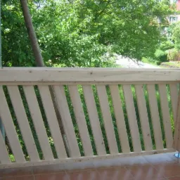 Surowa, drewniana balustrada balkonowa z ukośnymi szczeblami, widok na zieleń ogrodu, brązowa terakota na podłodze.