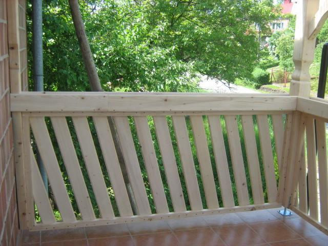 Surowa, drewniana balustrada balkonowa z ukośnymi szczeblami, widok na zieleń ogrodu, brązowa terakota na podłodze.