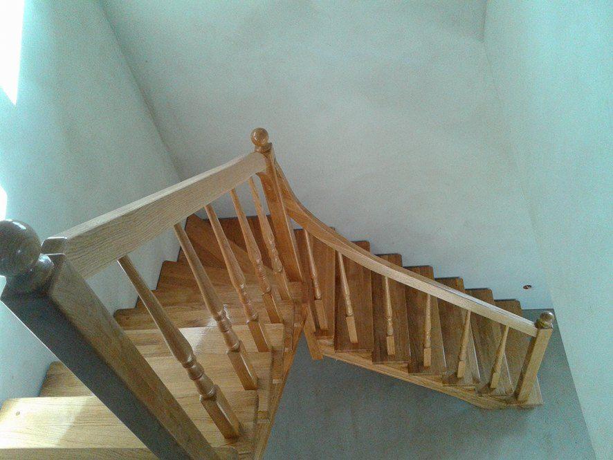 Widok z góry na drewniane schody z jasną, drewnianą balustradą i poręczą zakończoną kulką, w tle biała ściana i sufit.